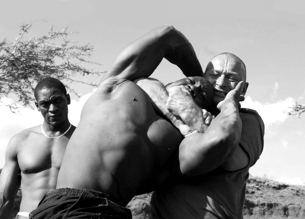 Mężczyzna w białej koszulce trzyma w chwycie innego mężczyznę bez koszulki. Obok stoi trzeci mężczyzna, obserwując sytuację. Tło to otwarte przestrzenie z drzewami.