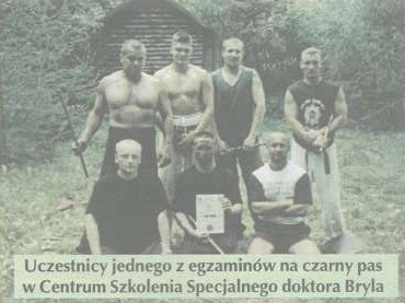 Mężczyźni stoją lub klęczą na trawie, trzymając certyfikaty i kije. W tle znajduje się drewniana chatka. Tekst: „Uczestnicy jednego z egzaminów na czarny pas w Centrum Szkolenia Specjalnego doktora Bryla.”