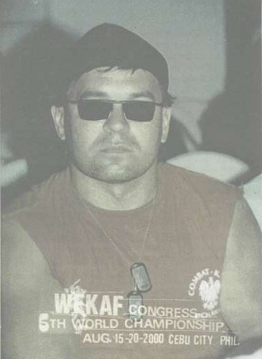 Jarosław Bielan w ciemnych okularach i czapce siedzi, patrząc na wprost. Nosi koszulkę z napisem: "WEKAF Congress 6th World Championship Aug. 15-20-2000 Cebu City, Phil." W tle widać rozmyte postacie.