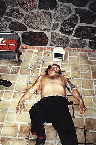 Jarosław Bielan leży na podłodze z odkrytym torsem, podłączony do urządzenia medycznego. Na ziemi widać też medyczne akcesoria. Tło stanowi ściana z kamienia.