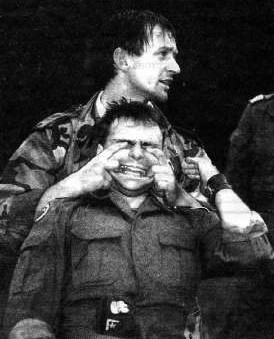 Dwóch mężczyzn w mundurach wojskowych rozgrywa scenkę; jeden stoi za drugim, trzymając go w uścisku. Tło jest ciemne, co tworzy dramatyczną atmosferę.