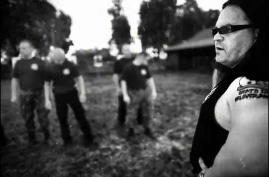 Jarosław Bielan w okularach przeciwsłonecznych stoi bokiem, pokazując tatuaż na ramieniu, na tle czterech osób w jednakowych koszulkach, na otwartym terenie z drzewami i budynkiem w oddali.