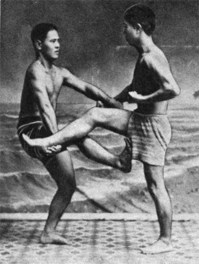 Dwóch młodych mężczyzn, w szortach, wykonuje technikę samoobrony w pozycji przy brzegiem morza. Jeden trzyma uniesioną nogę drugiego, tworząc dynamiczną scenę.