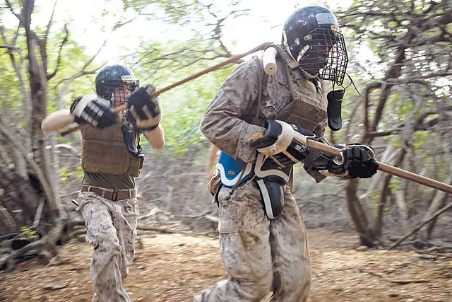 Dwaj żołnierze w ochronnych hełmach i mundurach trenują walkę kijami w lesie, biegnąc pomiędzy drzewami. Otoczenie jest zalesione, ziemia pokryta liśćmi.