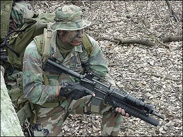 Żołnierz w pełnym umundurowaniu kuca, trzymając karabin, na tle lasu z opadłymi liśćmi i gołymi gałęziami. Całość tworzy atmosferę leśnej operacji wojskowej.