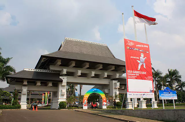 Brama w tradycyjnym indonezyjskim stylu stoi przed dużym plakatem z napisem „SEA GAMES XXVI Indonesia 2011”, w otoczeniu palm i flag.