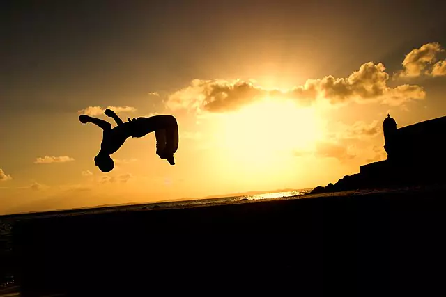 Sylwetka osoby wykonującej salto w tył na tle zachodzącego słońca. W tle widać słabo zarysowane fale oceanu i zarysy budynku.