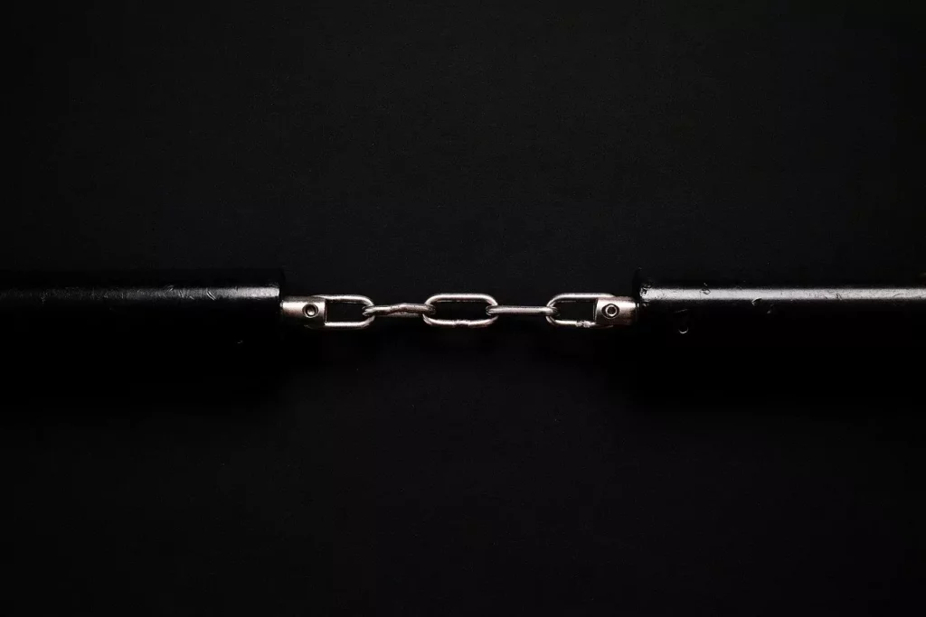 Dwie metalowe liny połączone krótkim łańcuchem, na czarnym tle. Centralny łańcuch trzyma oba końce razem, tworząc symetryczną, minimalistyczną kompozycję.