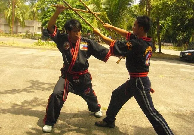 Dwóch mężczyzn ćwiczy walkę kijami na betonowej powierzchni, ubrani w tradycyjne stroje sztuk walki, otoczeni palmami i drzewami.