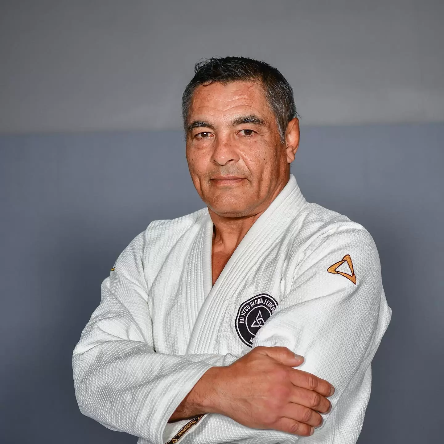 Mężczyzna w białym kimonie stoi z rękoma skrzyżowanymi na piersi, patrząc wprost. Na kimonie widnieje naszywka "Jiu-Jitsu Global Federation". Tło jest jednolicie szare.