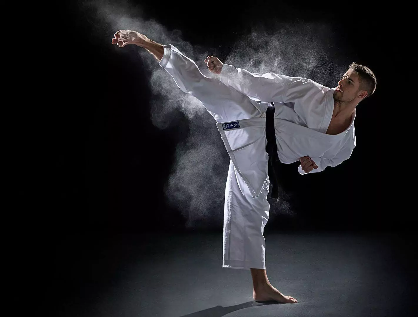 Mężczyzna w białym kimonie wykonuje wysokie kopnięcie w powietrzu w zadymionym otoczeniu na czarnym tle. Jego czarny pas podkreśla zaawansowanie w sztukach walki.