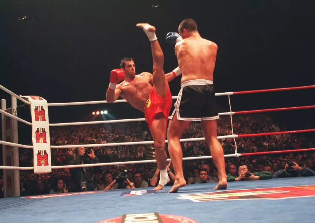 Dwóch zawodników walczy na ringu. Jeden wykonuje wysoki kopnięcie, drugi stoi w obronnej pozycji. W tle siedzą widzowie, oświetleni reflektorami.
