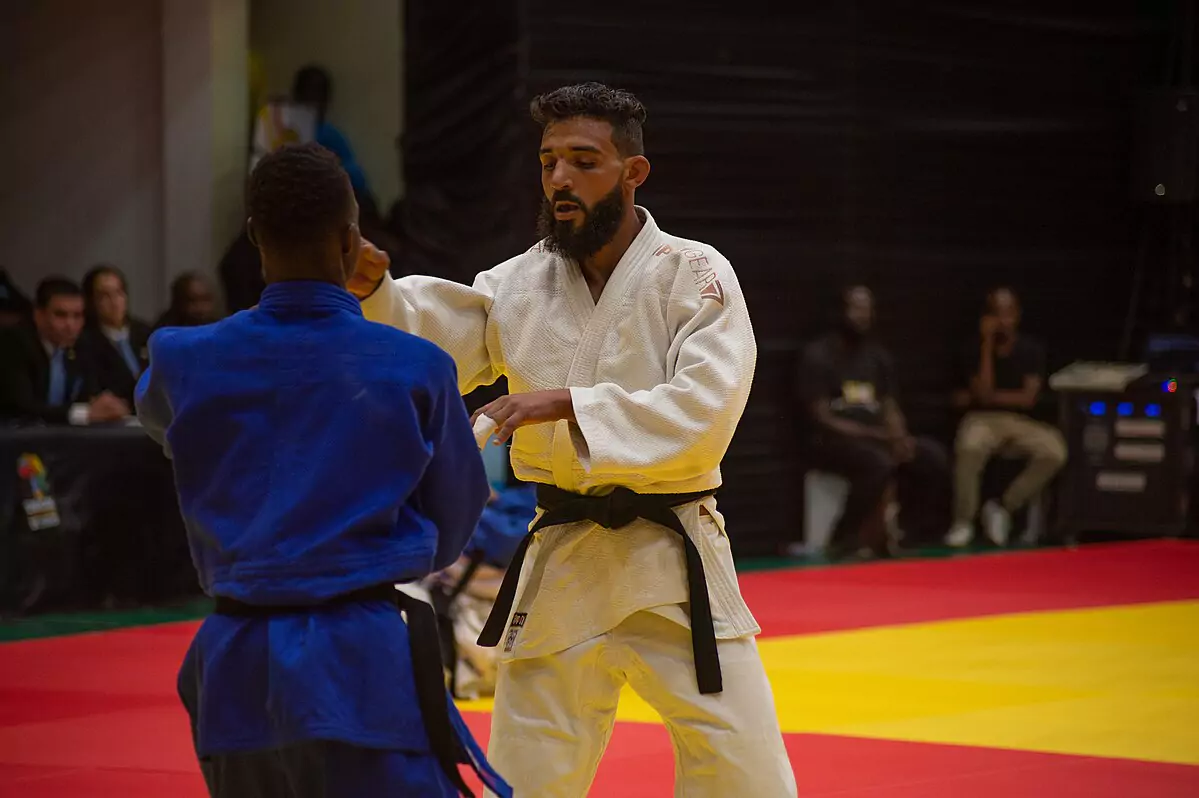 Zawodnik w białym judogi wykonuje technikę judo na przeciwniku w niebieskim stroju. Otoczony jest kolorową matą i widownią w tle.