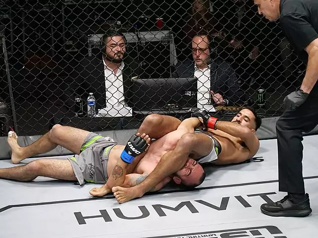 Dwóch zawodników walczy w oktagonie UFC; jeden usiłuje wykonać dźwignię na rękę przeciwnika. W tle za siatką siedzą sędziowie przy komputerach. Na podłodze widoczne jest logo „HUMVIT.”