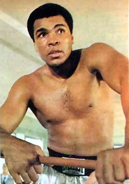 Muhammad Ali trenuje, opierając się o linę bokserskiego ringu, intensywnie patrząc w bok. Sportowiec jest w otoczeniu sali treningowej, z jasnym oświetleniem. Brak tekstu.