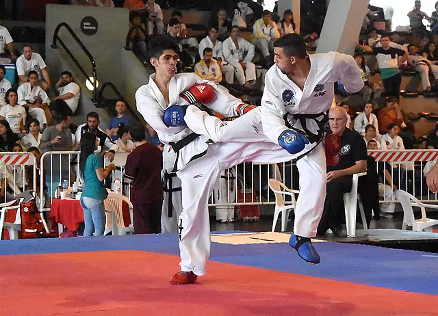 Dwóch zawodników taekwondo walczy na macie, wymieniając kopnięcia. W tle widać siedzących widzów i sędziów. Zawodnicy mają na sobie białe uniformy i kolorowe ochraniacze.