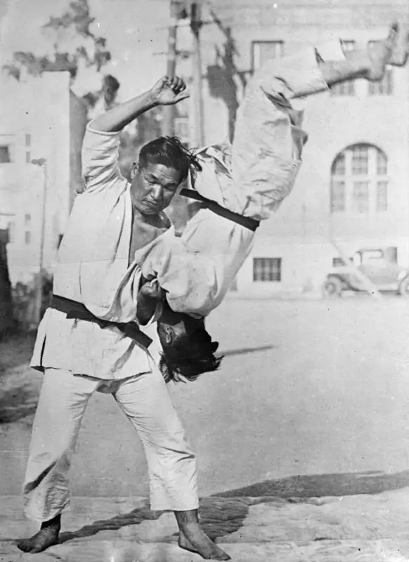 Dwóch mężczyzn w judogach wykonuje rzut judo na ulicy; jeden podnosi drugiego do góry nogami. W tle widoczne są budynki i zaparkowane samochody.