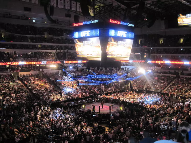 Ośmiokątna klatka, centrum uwagi wypełnionej areny, miejsca pełne widzów. Na wyświetlaczu widnieje jasno podświetlone logo "UFC", a nad nim napis "American Airlines Center".