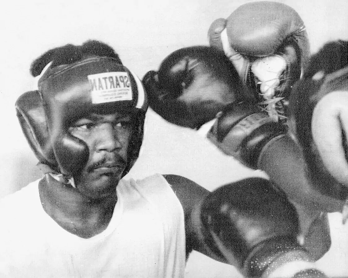 George Foreman w kasku z napisem "SPARTAN" blokuje cios w ringu. Ubrany w biały podkoszulek, skupiony na treningu, otoczony ramionami w rękawicach bokserskich.