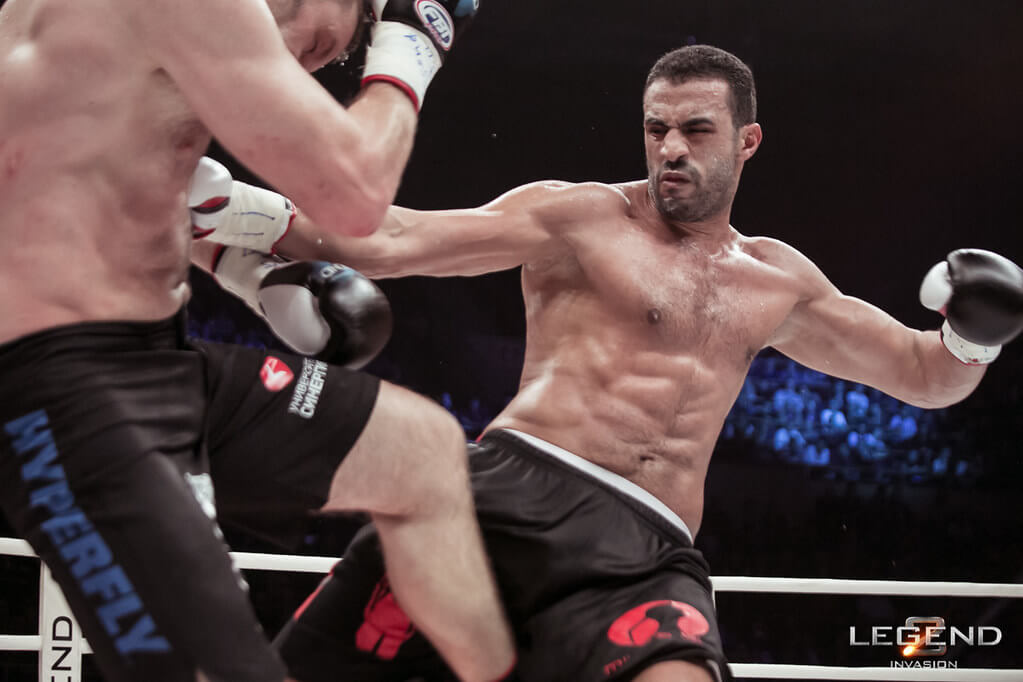 Dwóch zawodników w rękawicach bokserskich walczy na ringu. Badr Hari uderza pięścią, celując w przeciwnika. W tle widoczna jest publiczność. Napis: "LEGEND INVASION".