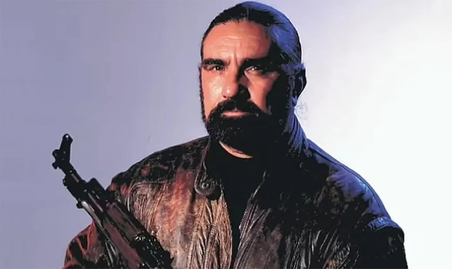 Richar Marcinko przytrzymuje karabin, patrząc intensywnie przed siebie. Ubrany w skórzaną kurtkę, stoi na jasnym tle.