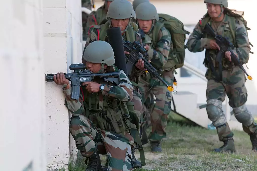 Żołnierze w pełnym oporządzeniu wojskowym, uzbrojeni, poruszają się w szyku przy ścianie budynku. Jeden z nich klęczy, celując karabinem, podczas gdy reszta idzie za nim. Grunt jest trawiasty.