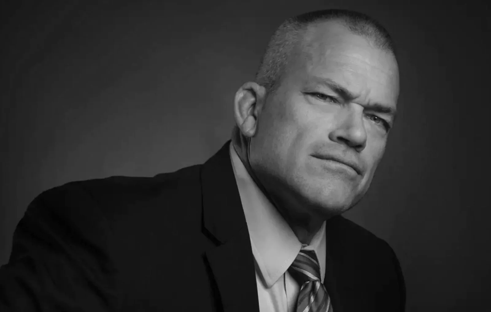 Mężczyzna w garniturze patrzy poważnie przed siebie, w lekko zacienionym, neutralnym tle.