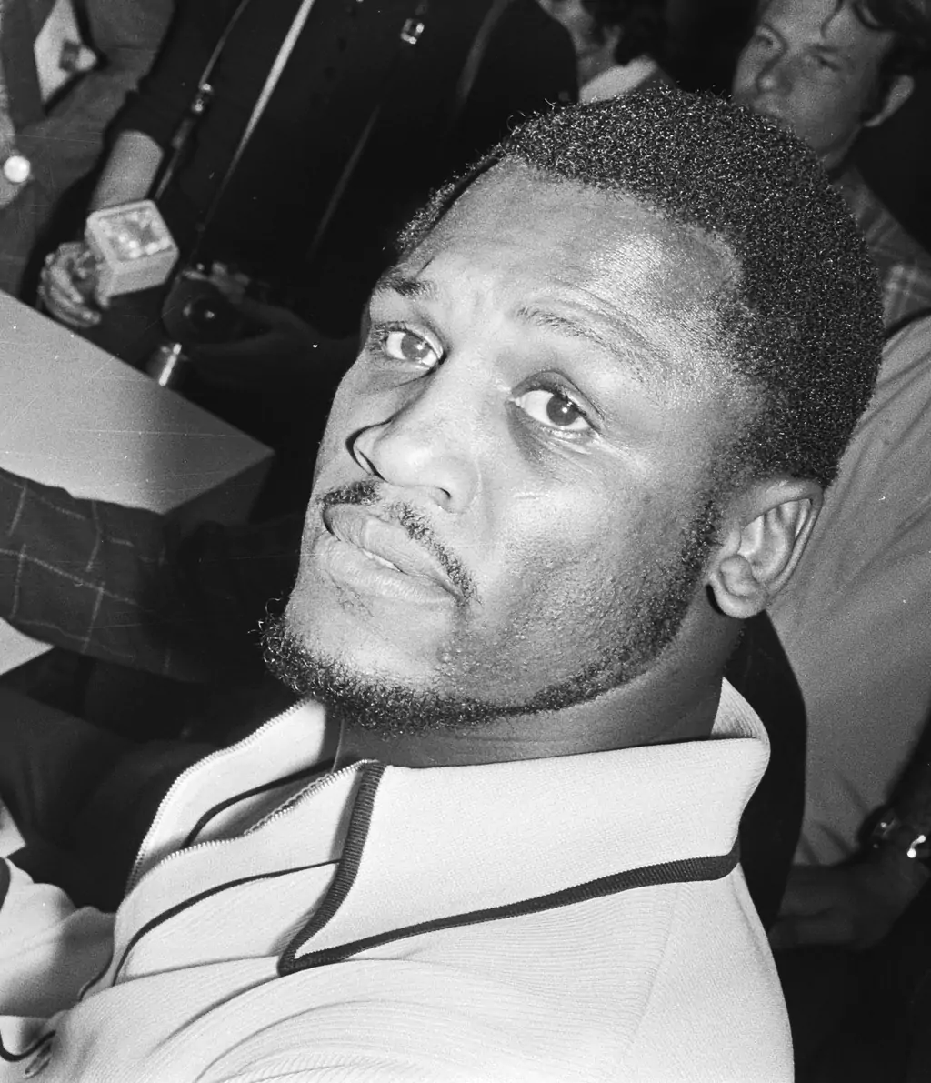 Mężczyzna w białej koszuli patrzy do góry. W tle widać grupę ludzi stojących blisko siebie. Atmosfera wydaje się być zatłoczona, prawdopodobnie w pomieszczeniu publicznym.