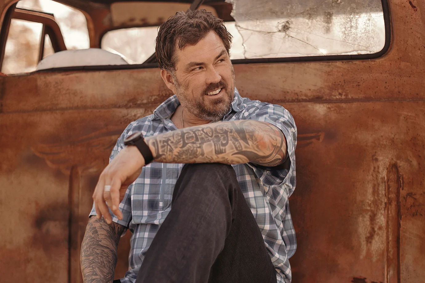 Mężczyzna siedzi, uśmiechając się, oparty o rdzewiejącą, stary samochód. Ma brodę, tatuaże na ramionach i koszulę w kratę. Za nim widać rozbity szkielet pojazdu.