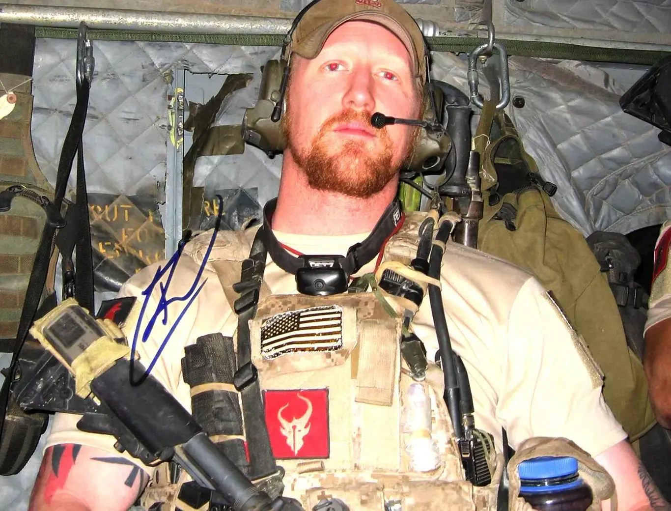 Mężczyzna w wojskowym wyposażeniu siedzi w wnętrzu pojazdu wojskowego. Ma na sobie hełm oraz kamizelkę ochronną z naszywką flagi USA. Podpis na obrazie: „K” (odręczna sygnatura).