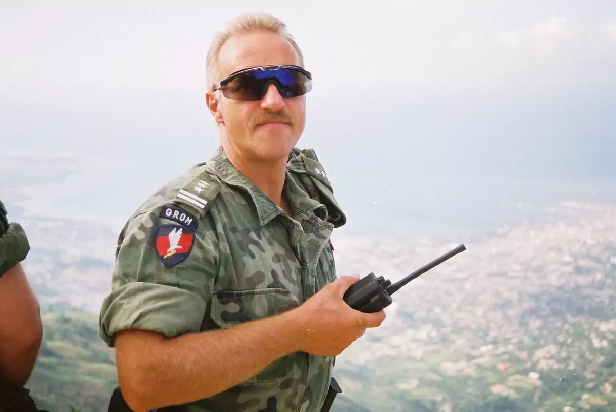 Mężczyzna w mundurze wojskowym, z plakietką „GROM”, trzyma krótkofalówkę. Znajduje się na wysokości, z widokiem na miasto w tle. Ma okulary przeciwsłoneczne i wąsy.