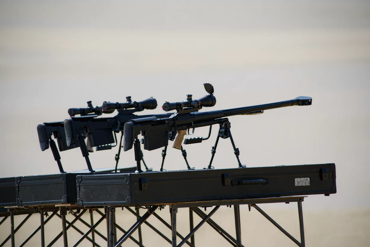 Karabiny snajperskie ustawione na stalowych skrzyniach, spoczywają na trójnogach, skierowane w prawą stronę. Tło jest jednolite, co kontrastuje z ciemnym kolorem broni.