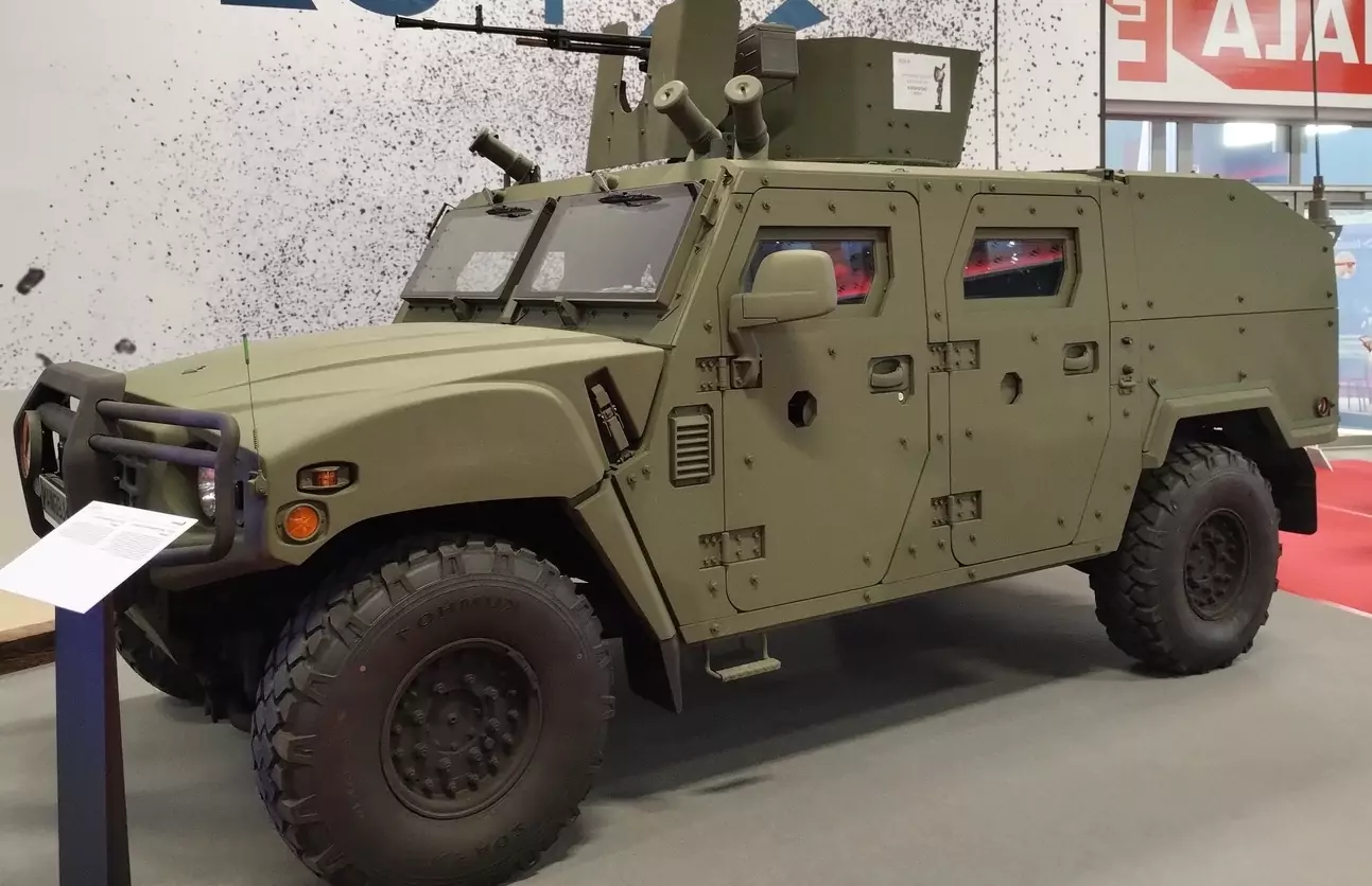Pojazd wojskowy o zielonym kolorze stoi na wystawie. Ma opancerzoną konstrukcję, duże terenowe opony i zamontowaną u góry broń. W tle widać częściowo widoczne logo i napis.