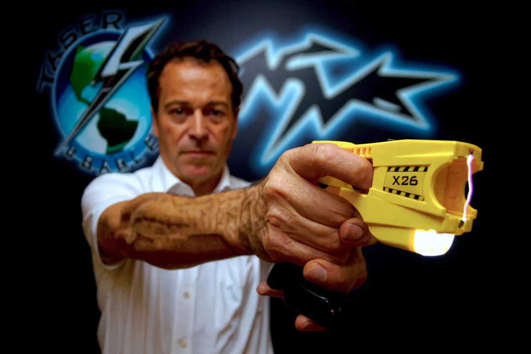 Mężczyzna trzyma żółty paralizator X26, mierząc nim w stronę kamery. W tle znajduje się logo z napisem „TASER”.