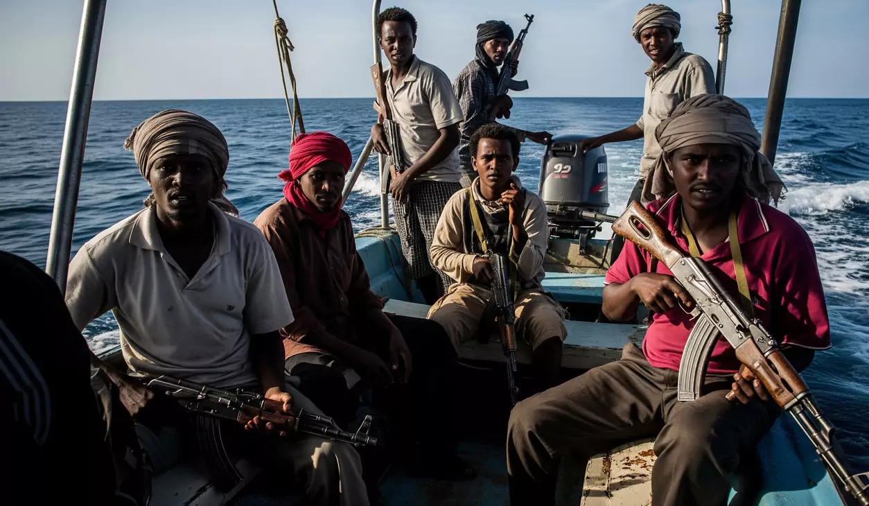 Grupa mężczyzn z bronią siedzi na niewielkiej łodzi na otwartym morzu. Łódź wyposażona w silnik zaburtowy płynie po spokojnej wodzie pod bezchmurnym niebem.