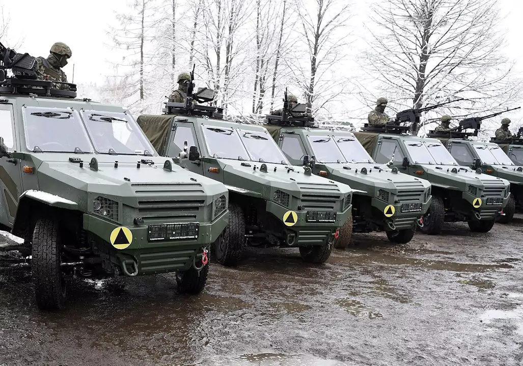 Opancerzone pojazdy wojskowe stoją w rzędzie na zaśnieżonej polanie, otoczone drzewami bez liści. Żołnierze siedzą w wieżyczkach pojazdów, przygotowani do działania.