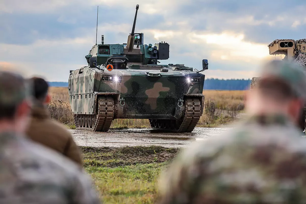 Pojazd wojskowy jedzie po wyboistej drodze; na horyzoncie widać pola i las. Dwóch żołnierzy stoi w rozmazaniu na pierwszym planie.