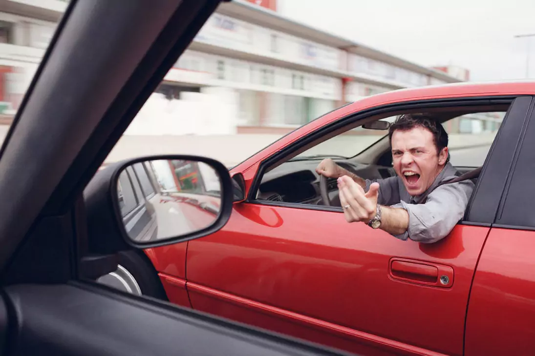 Kierowca wychyla się przez okno czerwonego samochodu, wykonując gniewny gest dłonią. Znajduje się na drodze, widoczne są inne pojazdy i budynki w tle.