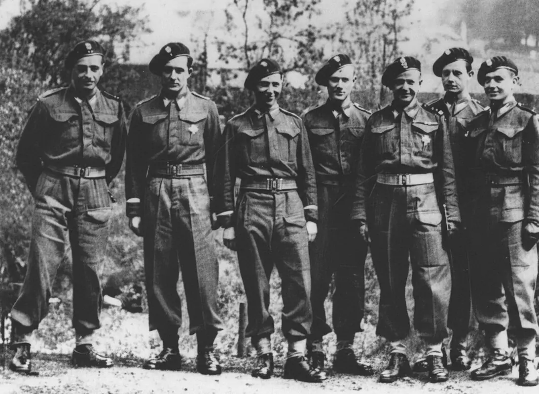 Żołnierze stoją w szeregu, ubrani w mundury wojskowe z beretami na głowach. Znajdują się na zewnątrz, na tle zarośli lub drzew.
