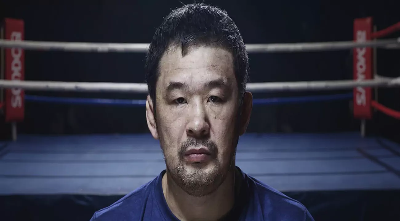 Mężczyzna patrzy w kamerę, stoi nieruchomo, z poważnym wyrazem twarzy. W tle widać ring bokserski z linami i czerwonymi słupkami.