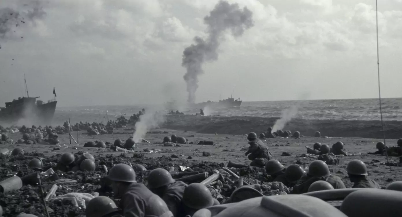 Żołnierze leżą na plaży, osłaniając się i przygotowując do ataku. W tle widoczne statki i dym unoszący się nad horyzontem, sugerujący bitwę morską.