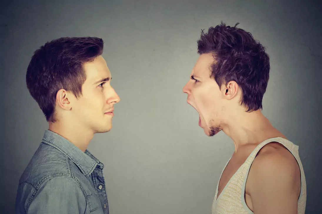 Dwóch mężczyzn stoi naprzeciwko siebie na neutralnym tle. Jeden z nich krzyczy, podczas gdy drugi spokojnie patrzy przed siebie, nie reagując na emocje pierwszego.