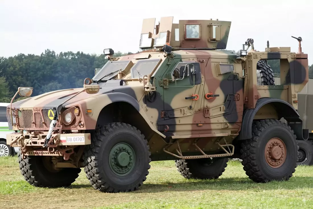 Pojazd wojskowy w kamuflażu stoi na trawie. Ma masywne koła i specjalne pancerze. W tle widoczne są drzewa i fragment innego pojazdu.
