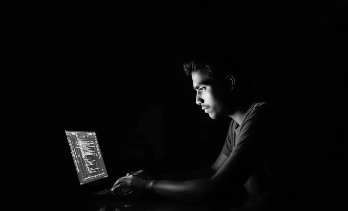 Mężczyzna pracuje przy laptopie, wpatruje się w ekran z kodem, w ciemnym pomieszczeniu z oświetleniem skupionym na jego twarzy.