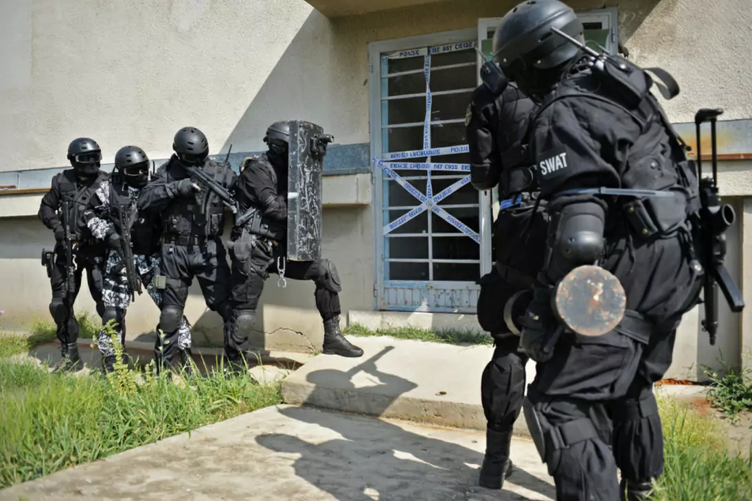 Funkcjonariusze jednostki SWAT w czarnych mundurach, uzbrojeni, wchodzą do budynku. Jeden niesie tarczę ochronną. Drzwi zabezpieczone taśmą z napisem "POLICE LINE DO NOT CROSS".