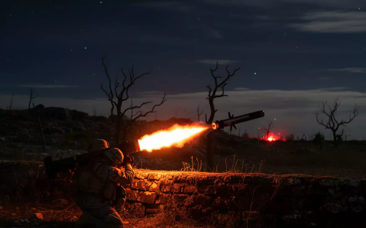 Żołnierz wystrzeliwuje pocisk z broni przeciwpancernej. Działa nocą pośród suchego, pustynnego terenu z martwymi drzewami w tle. Atmosfera jest wzmacniana przez jasny, ognisty wystrzał.