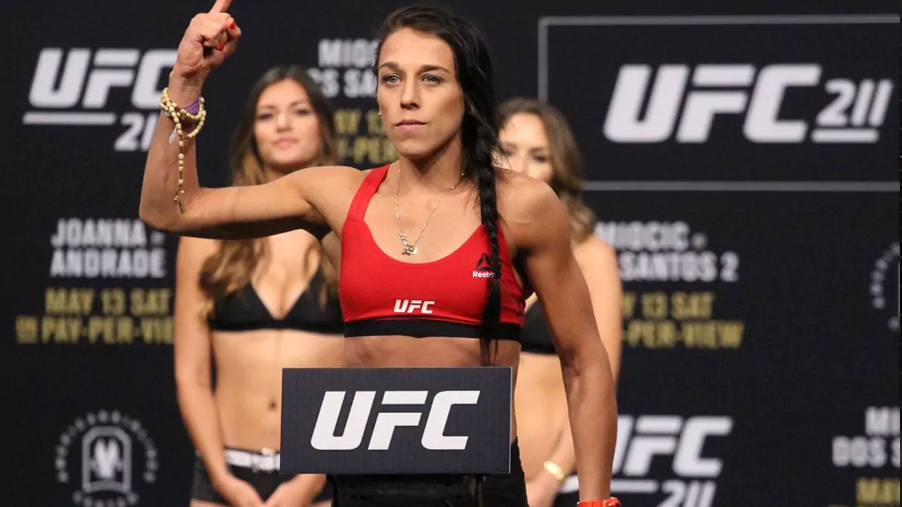 Kobieta w sportowym stroju UFC unosi pięść na tle banerów promujących galę UFC 211. W tle widać inne osoby stojące na scenie.