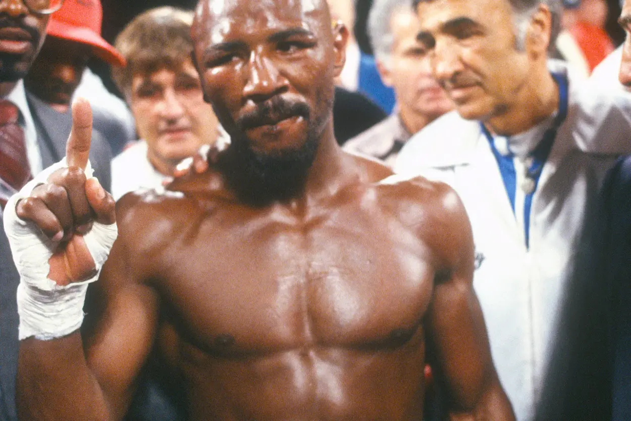 Mężczyzna z gołą klatką piersiową unosi zaciśniętą dłoń z wyprostowanym palcem wskazującym. Otaczają go ludzie w tle, a atmosfera jest napięta.