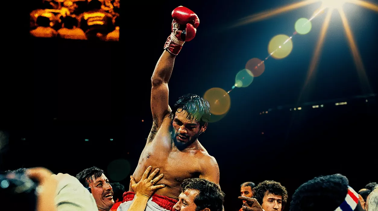 Bokser z uniesioną ręką w czerwonej rękawicy, wyrażający triumf. Wokół niego grupa ludzi podtrzymuje go. Tło jest ciemne z jasnym światłem reflektora.