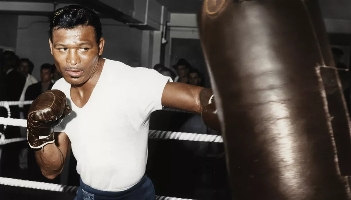 Bokser uderza worek treningowy na siłowni, koncentrując się na swoim zadaniu. Wokół znajdują się liny ringu, a w tle widać zarys kilku osób obserwujących ćwiczenia.